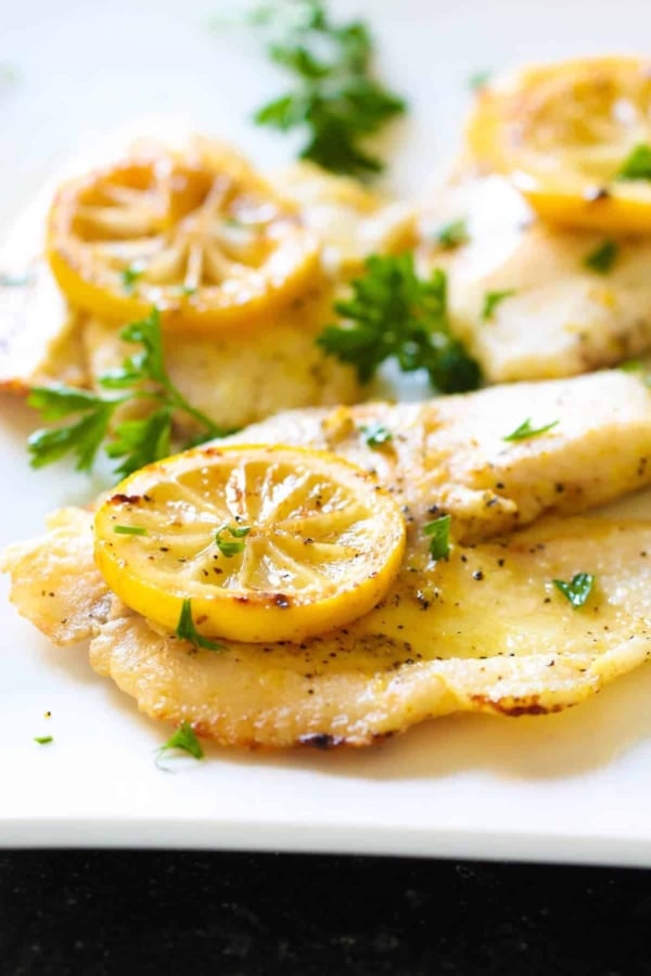 Cooked fish fillets garnished with slices of lemon and chopped parsley on a white plate.