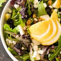 A bowl of mixed green salad topped with sliced pear, dried cranberries, walnuts, and crumbled cheese.