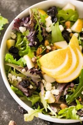 A bowl of mixed green salad topped with sliced pear, dried cranberries, walnuts, and crumbled cheese.