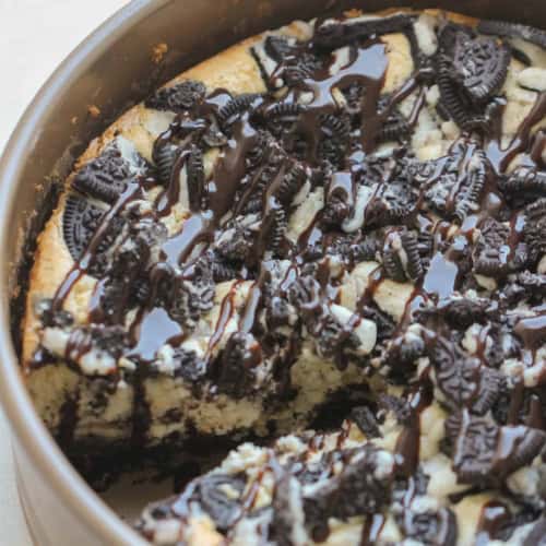 A cheesecake in a round pan topped with crushed cookies and chocolate drizzle, with a slice missing.