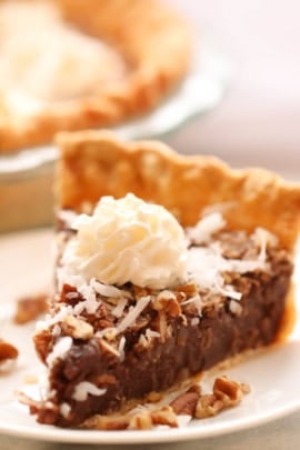 A slice of chocolate pie topped with whipped cream, shredded coconut, and chopped nuts on a white plate. Another pie is visible in the background.