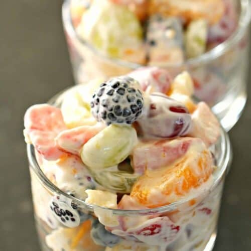 Two glass cups filled with a creamy fruit salad containing grapes, blackberries, orange segments, and other assorted fruits.