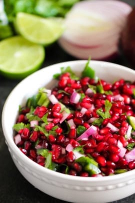 A bowl of pomegranate salsa with chopped red onions and cilantro, garnished with lime slices in the background.