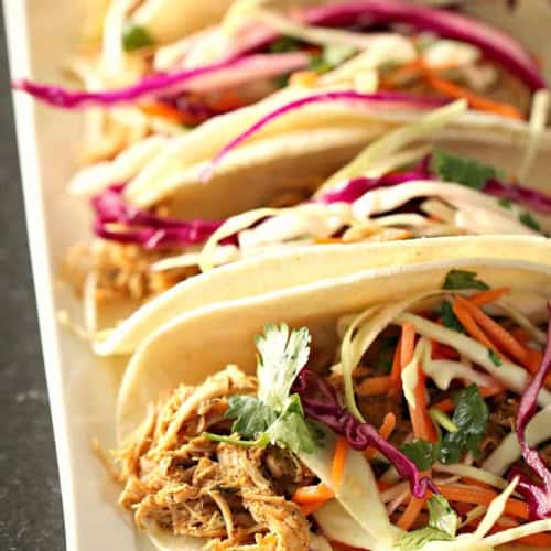 A row of soft shell tacos filled with shredded meat, shredded cabbage, carrots, and fresh cilantro, arranged on a white rectangular plate.