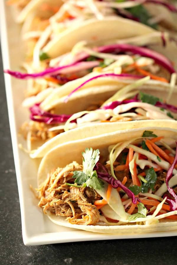 A row of soft shell tacos filled with shredded meat, shredded cabbage, carrots, and fresh cilantro, arranged on a white rectangular plate.