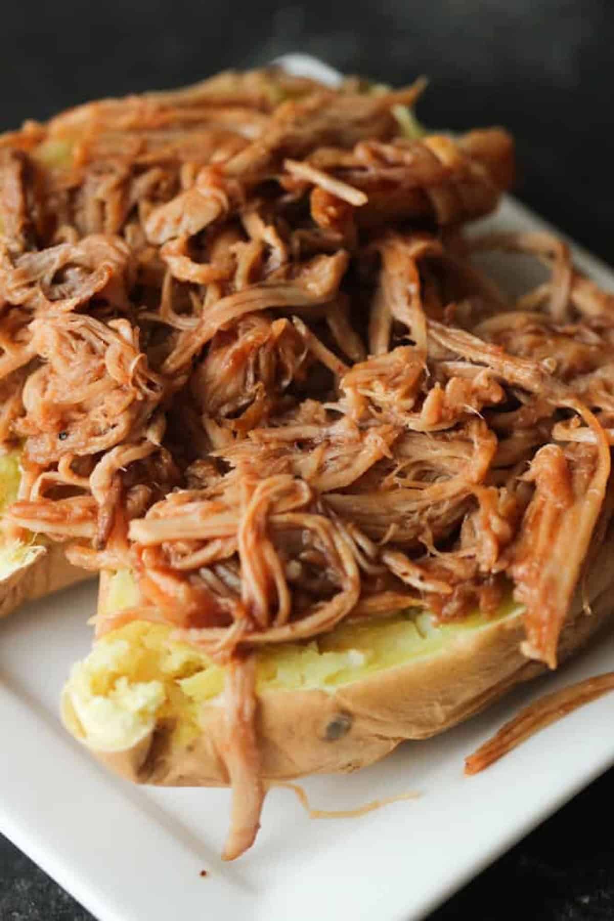 A baked potato topped with shredded barbeque pulled pork on a white plate.