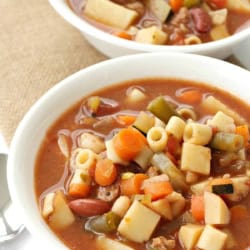 Two bowls of vegetable and pasta soup on a burlap cloth. The soup contains pieces of pasta, carrots, green beans, and other assorted vegetables.
