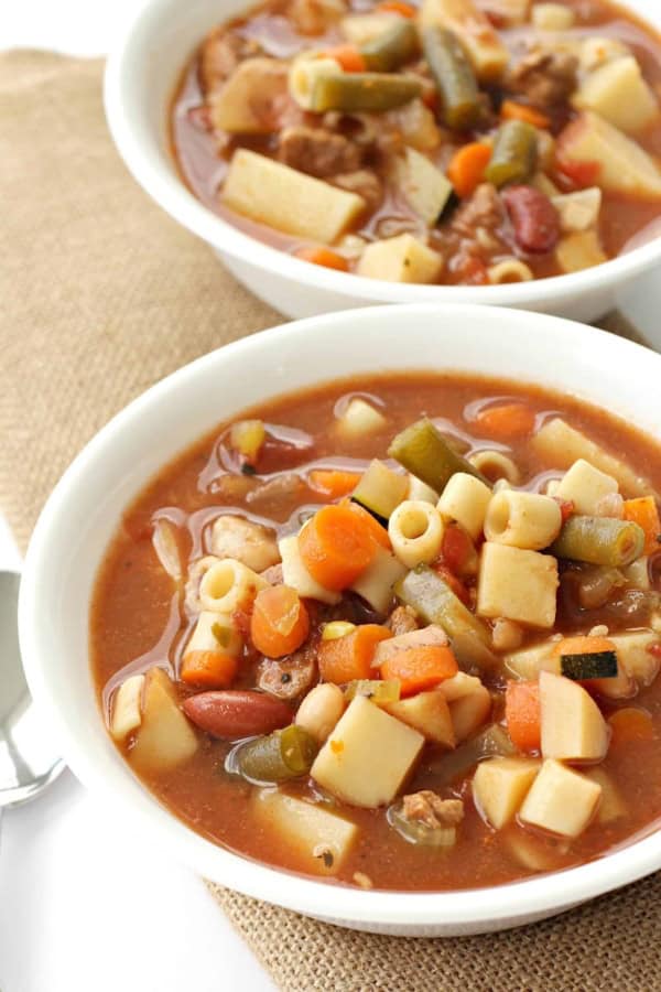 Two bowls of vegetable and pasta soup on a burlap cloth. The soup contains pieces of pasta, carrots, green beans, and other assorted vegetables.