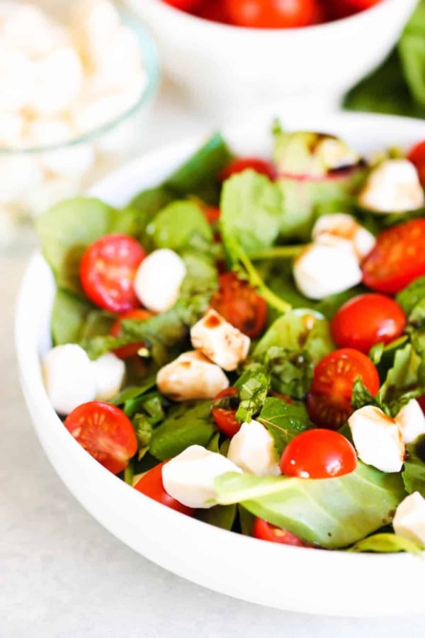 A white bowl filled with a salad of fresh spinach leaves, cherry tomatoes, mozzarella balls, and a drizzle of balsamic dressing.