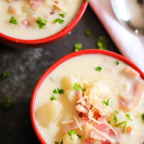 Two bowls of creamy soup garnished with chopped parsley, shredded cheese, and chunks of bacon, placed on a dark surface next to a white napkin and spoons.