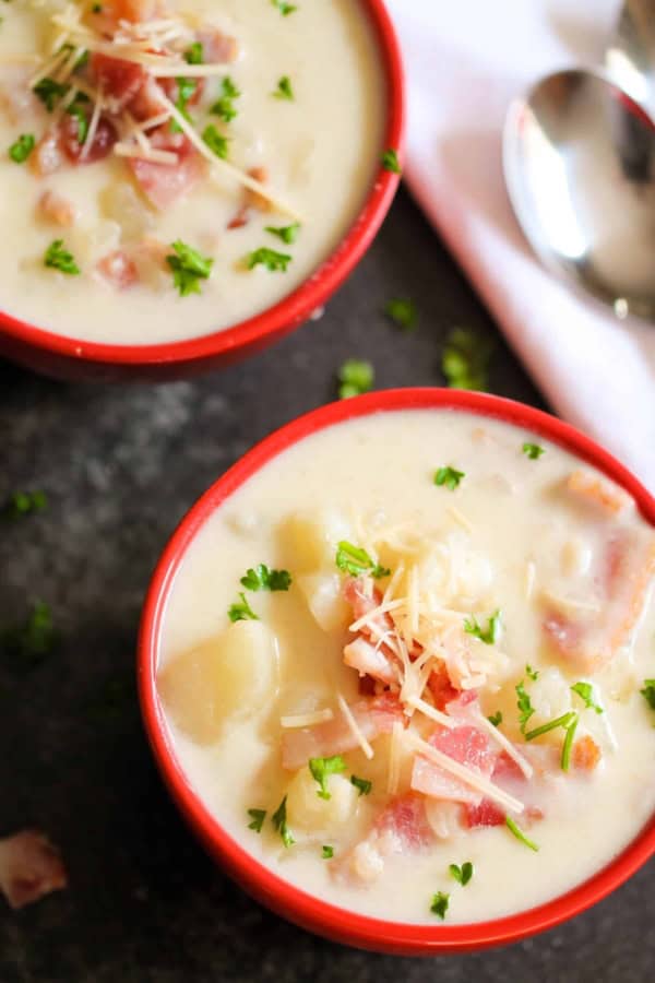 Two bowls of creamy soup garnished with chopped parsley, shredded cheese, and chunks of bacon, placed on a dark surface next to a white napkin and spoons.