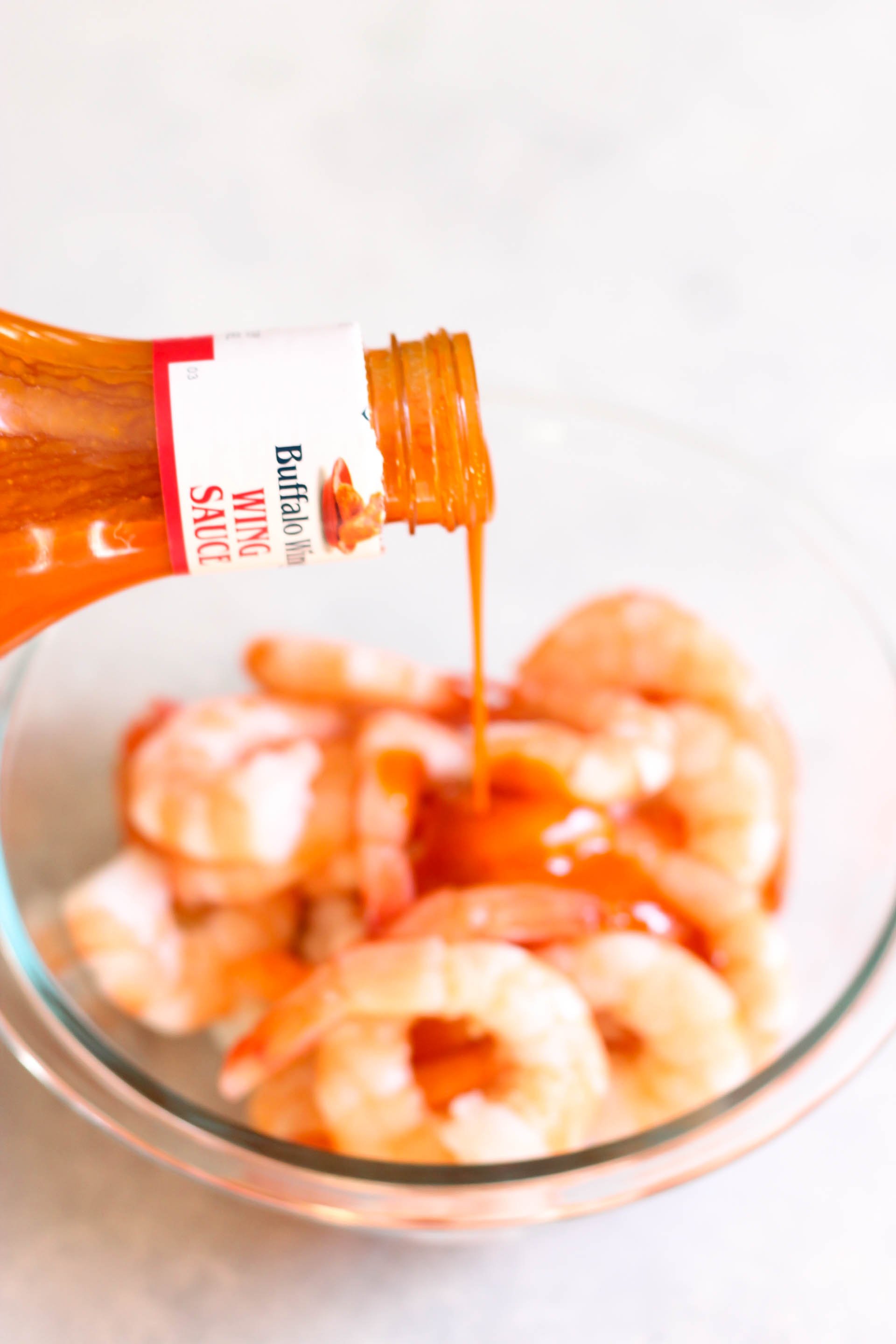 Raw shrimp in a mixing bowl with wing sauce being poured on top