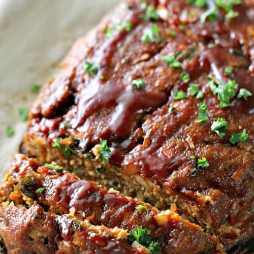 Sliced meatloaf topped with a glossy sauce and sprinkled with chopped parsley, resting on parchment paper.