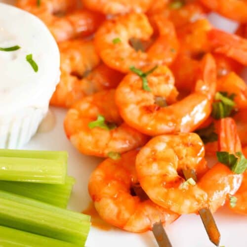 Cooked shrimp skewers garnished with herbs, served with celery sticks and a side of white dipping sauce on a white plate.