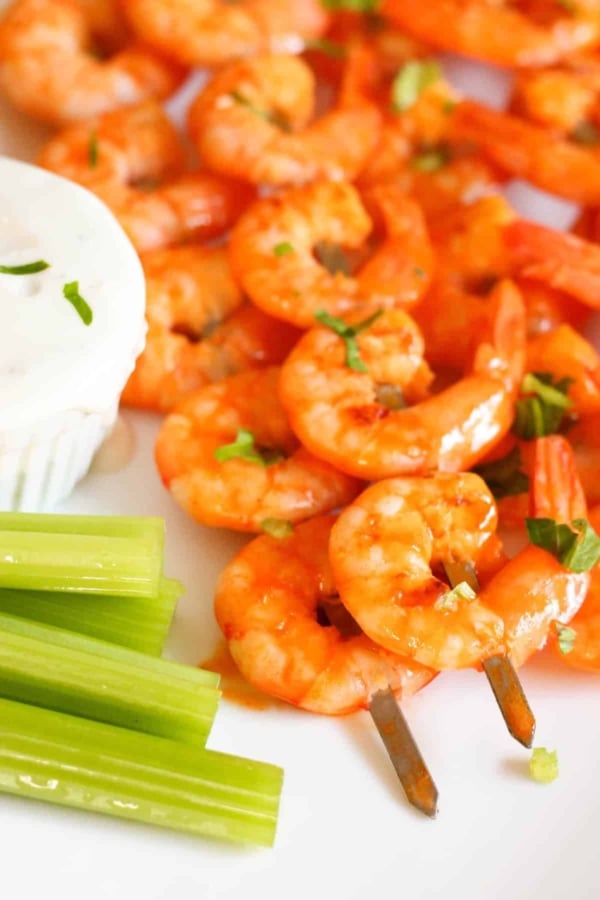 Cooked shrimp skewers garnished with herbs, served with celery sticks and a side of white dipping sauce on a white plate.