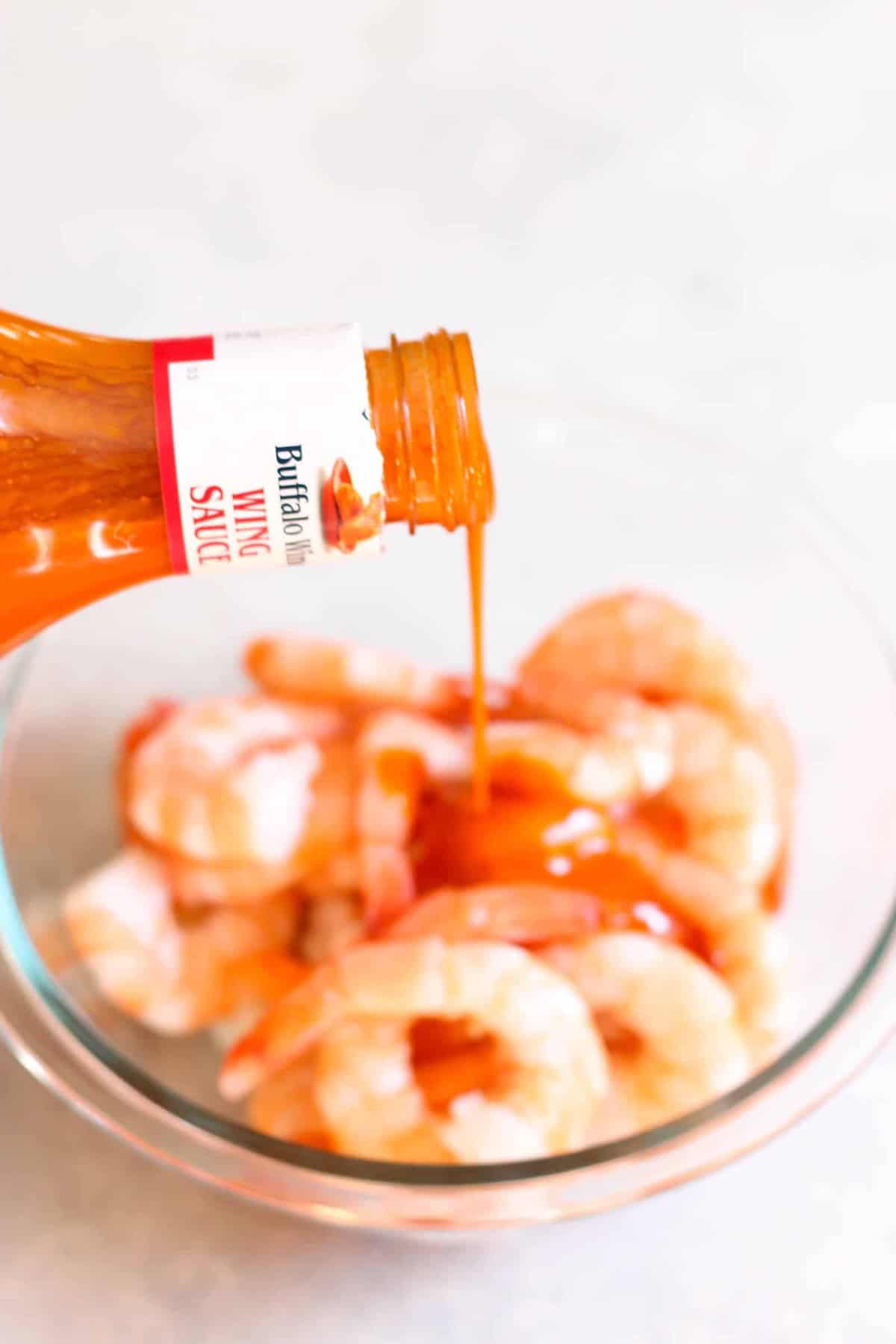 A bottle of buffalo wing sauce being poured over a bowl of raw shrimp.