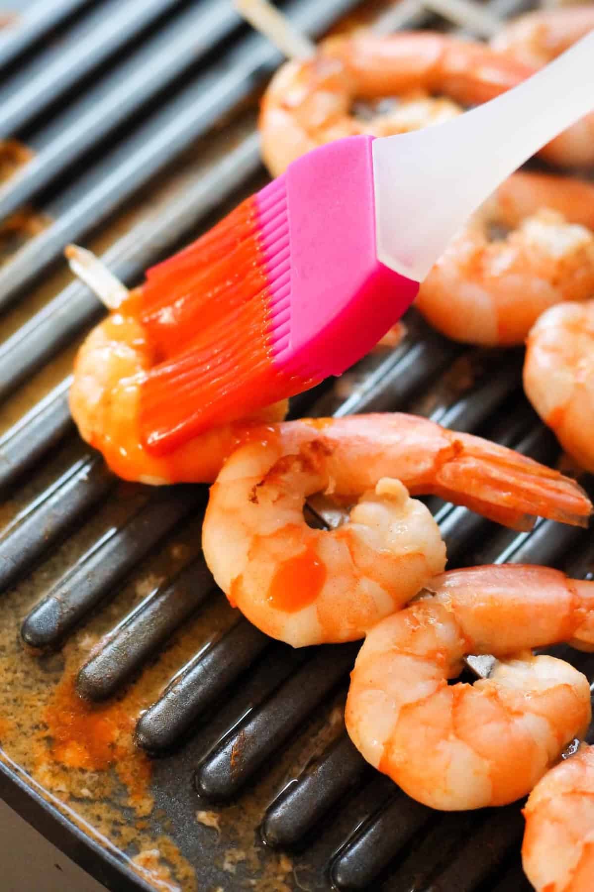 A silicone brush applies sauce to shrimp skewers cooking on a grill.