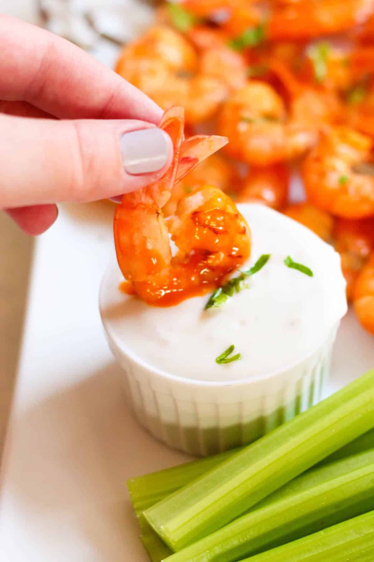 A hand dips a cooked shrimp into a small ramekin of white dipping sauce, with celery sticks and more shrimp visible in the background.