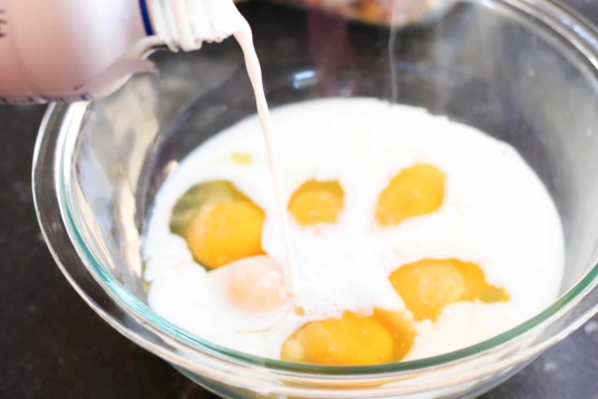 Milk being poured into a glass bowl with five raw eggs, preparing a mixture.