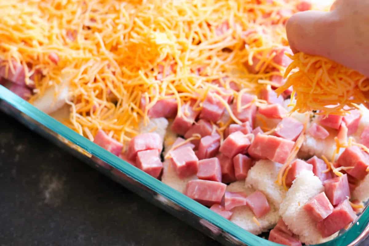 A hand sprinkles shredded cheese over a casserole dish filled with diced ham and cubed bread.