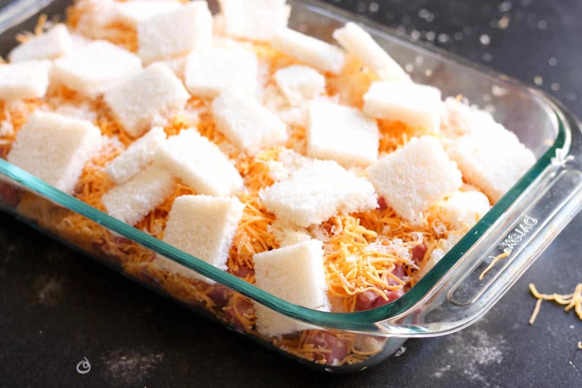 Glass baking dish with layers of shredded cheese, cubed bread, and visible ingredients on a dark surface.