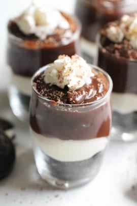 Close-up of layered dessert cups filled with chocolate pudding, a white cream layer, and topped with whipped cream and chocolate shavings.