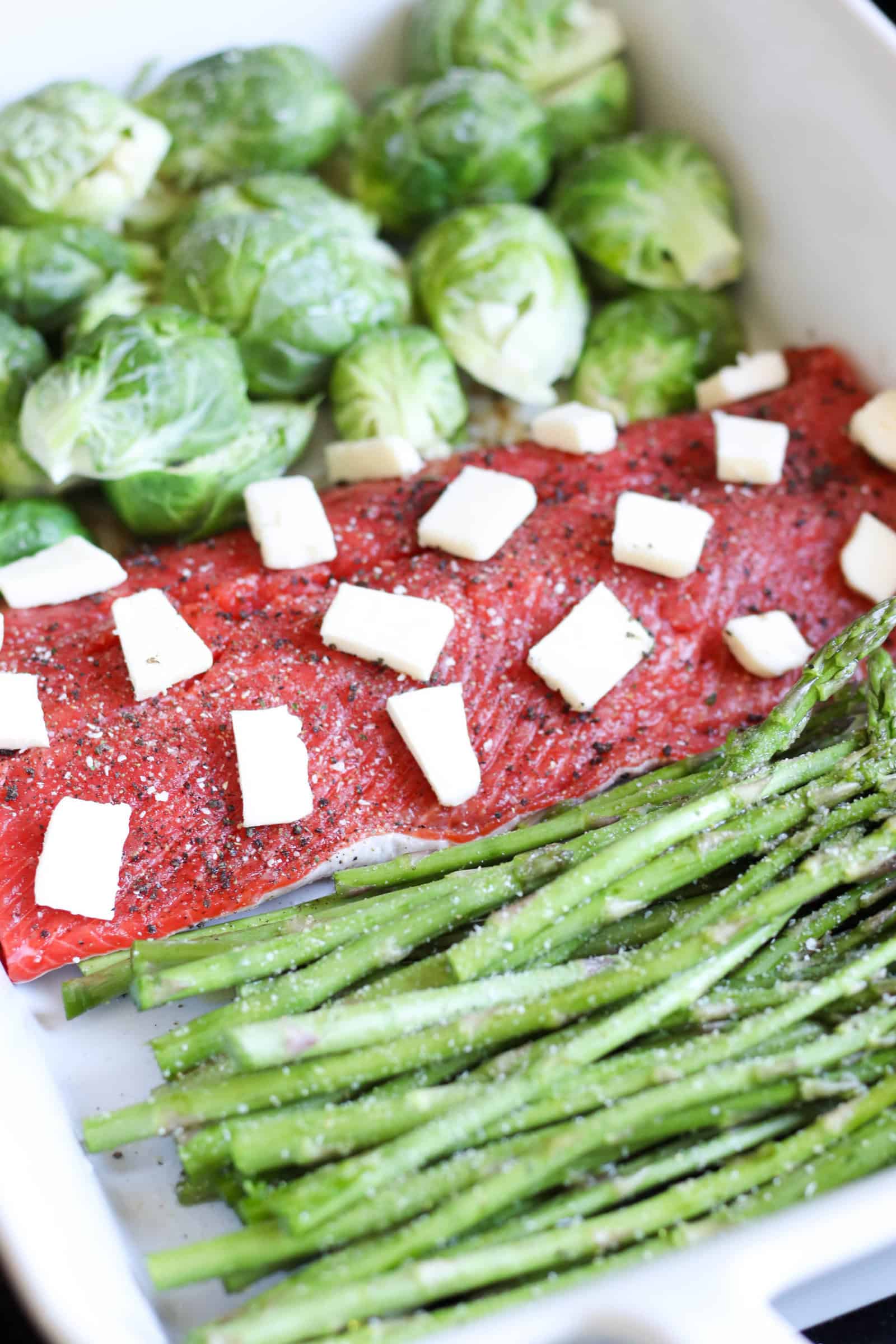 Raw salmon topped with butter slices on a pan with brussels sprouts and asparagus