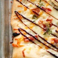 Rectangular flatbread topped with melted cheese, chopped herbs, diced meat, and drizzled with a dark sauce on a baking sheet.