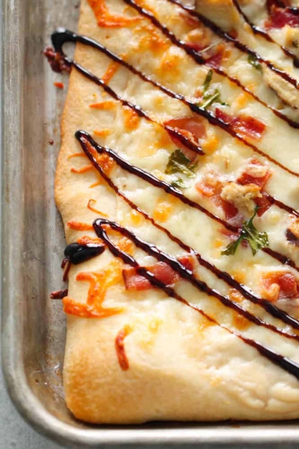 Rectangular flatbread topped with melted cheese, chopped herbs, diced meat, and drizzled with a dark sauce on a baking sheet.