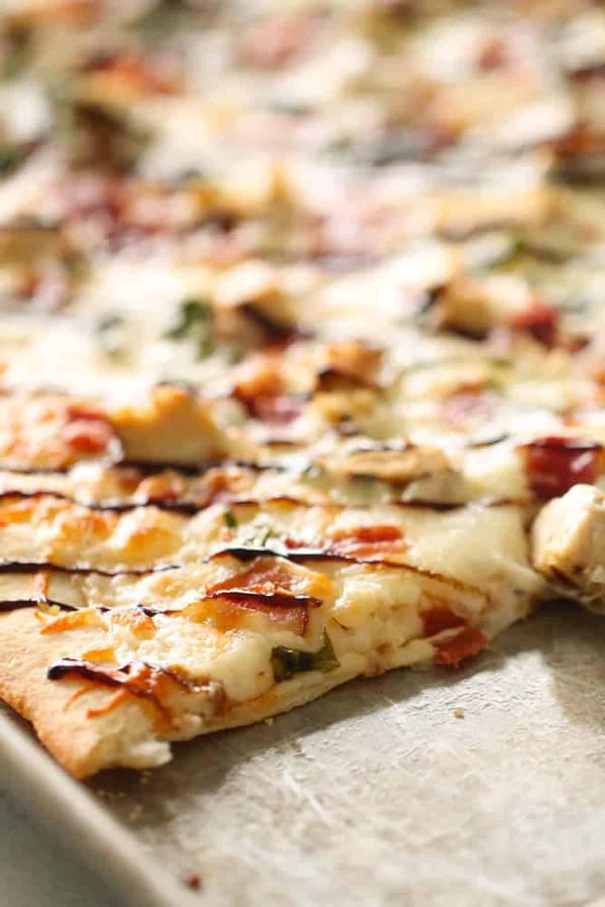 Close-up of a rectangular flatbread pizza with melted cheese, bacon, chicken, herbs, and a drizzle of balsamic glaze on a baking sheet.