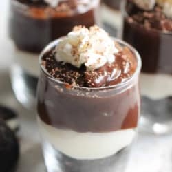 A glass filled with layered dessert consisting of chocolate pudding, a white creamy layer, crushed cookie base, and topped with whipped cream and chocolate shavings.