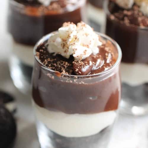 A glass filled with layered dessert consisting of chocolate pudding, a white creamy layer, crushed cookie base, and topped with whipped cream and chocolate shavings.