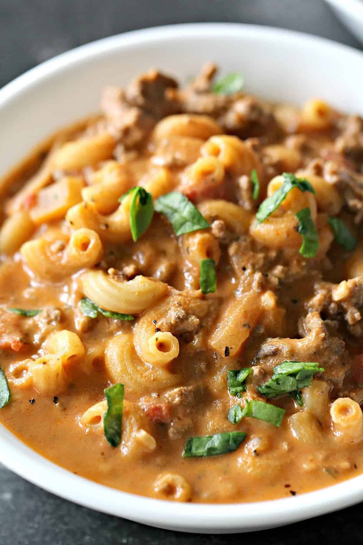 A bowl of creamy pasta with ground meat, elbow macaroni, tomato sauce, and fresh basil garnish.
