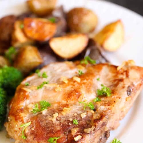 A cooked pork chop garnished with herbs, served with roasted potatoes and broccoli on a white plate.