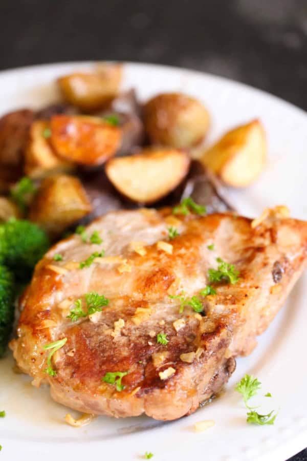 A cooked pork chop garnished with herbs, served with roasted potatoes and broccoli on a white plate.