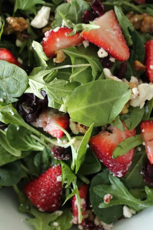 Close-up of a fresh salad with spinach, arugula, sliced strawberries, crumbled feta cheese, walnuts, and possibly dried cranberries.