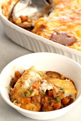 A serving of shepherd's pie in a white bowl with a larger casserole dish of shepherd's pie in the background.