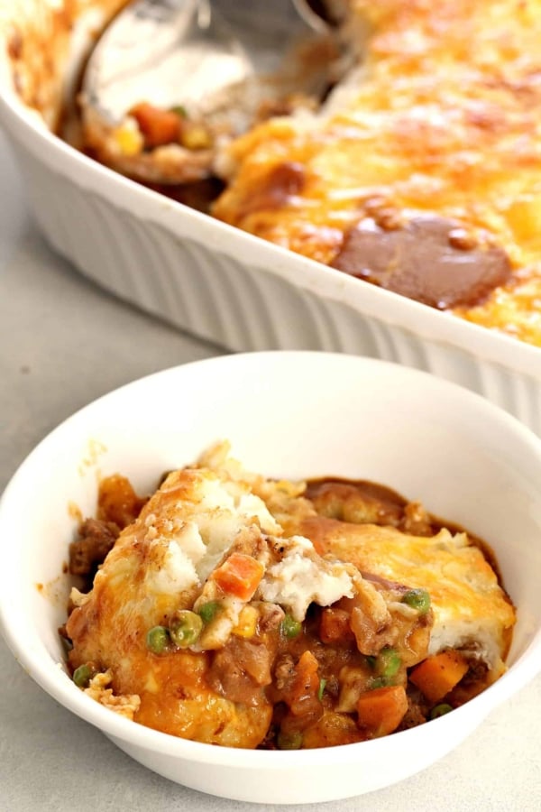 A serving of shepherd's pie in a white bowl with a larger casserole dish of shepherd's pie in the background.