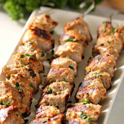 Three skewers of grilled meat garnished with herbs are arranged on a rectangular white plate, with parsley and an onion in the background.