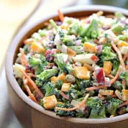 A wooden bowl filled with chopped broccoli, carrots, cheese cubes, red onion, and diced apples mixed in a creamy dressing.