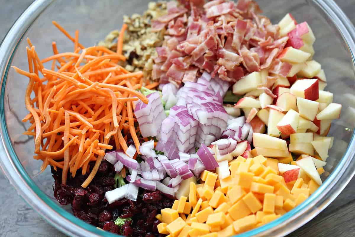 A glass bowl containing chopped apples, cheddar cheese cubes, red onions, shredded carrots, dried cranberries, walnuts, and cooked bacon pieces arranged in separate piles.