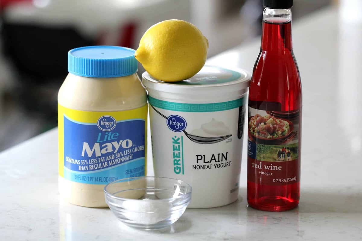A jar of lite mayonnaise, a tub of plain Greek yogurt, a lemon, a bottle of red wine vinegar, and a small bowl of salt on a white countertop.