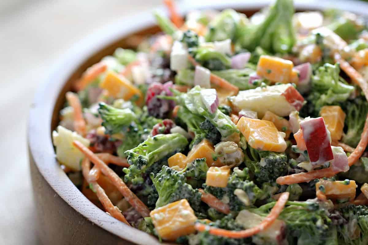 A wooden bowl filled with a broccoli salad containing chopped broccoli, shredded carrots, diced cheese, red onions, and a creamy dressing.