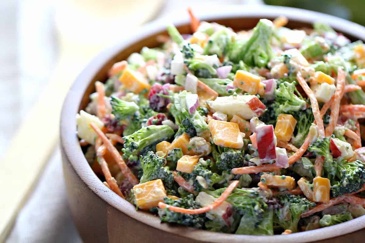 A wooden bowl filled with a broccoli salad containing chopped broccoli, shredded carrots, diced cheese, red onions, and a creamy dressing.