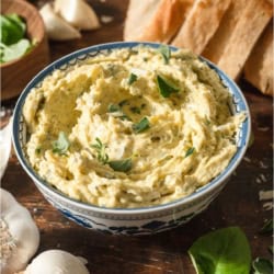 A bowl of Garlic Butter with slices of bread in the background and garlic cloves.