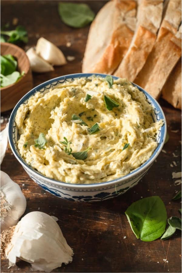 A bowl of Garlic Butter with slices of bread in the background and garlic cloves.