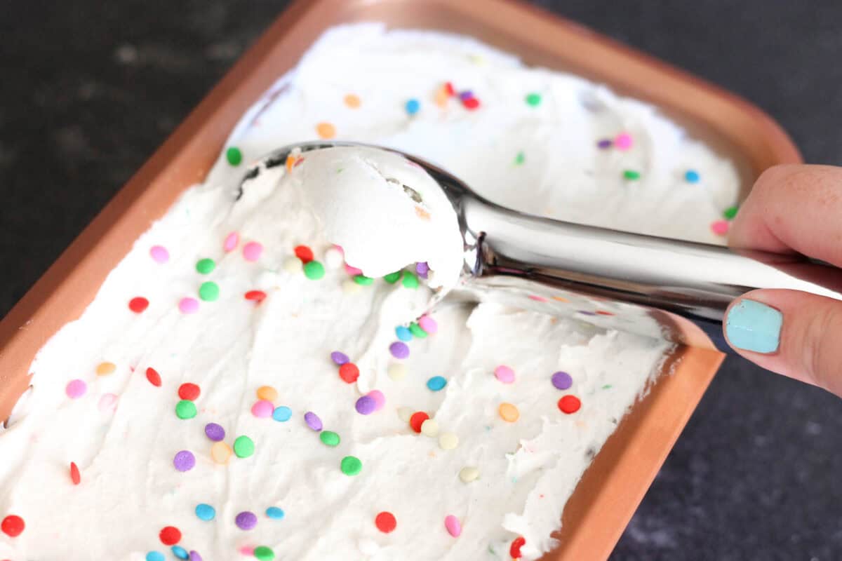 An ice cream scoop, scooping No-Churn Birthday Cake Protein Ice Cream from a pan.