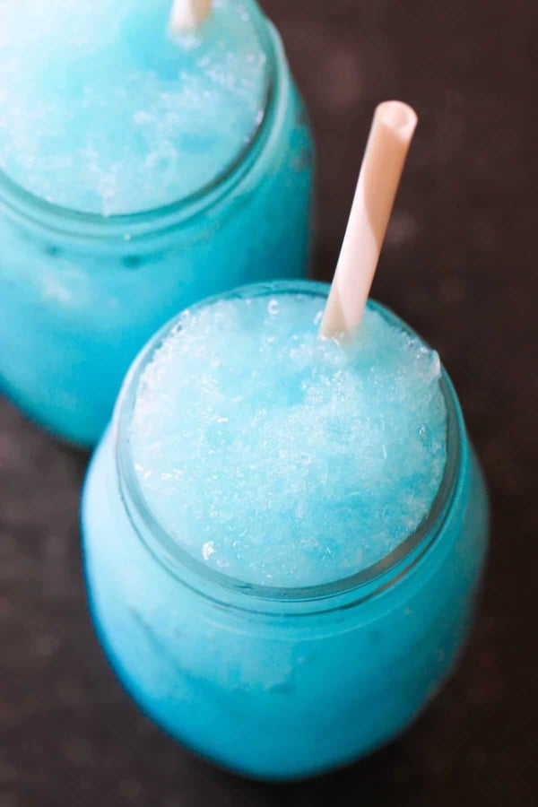 Two glass jars filled with bright blue slushy drinks, each with a white straw, viewed from above on a dark surface.