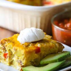Close-up of a slice of savory casserole topped with sour cream on a plate, accompanied by avocado slices. A dish of salsa is visible in the background.