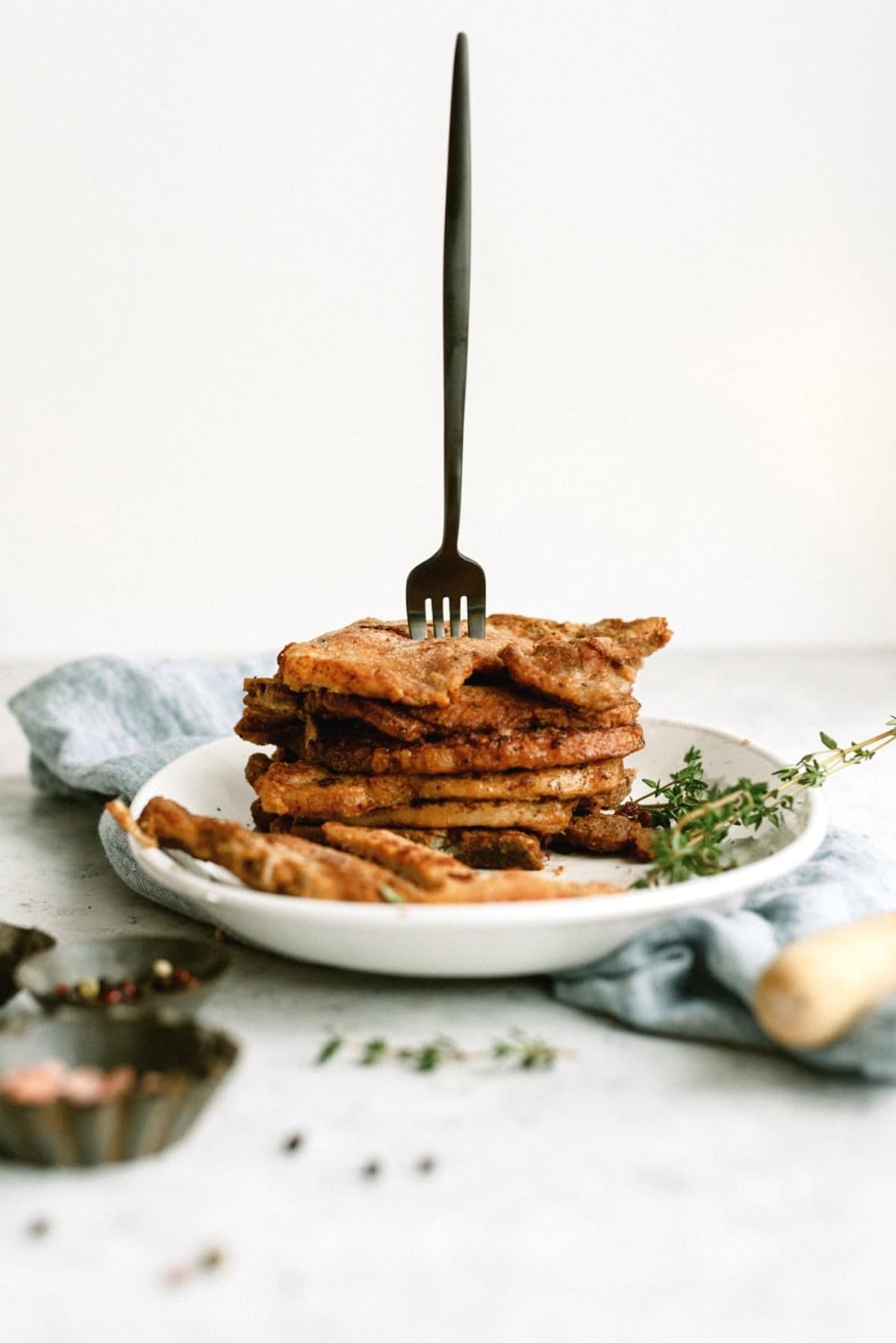 A stack of pork chops on a white plate with a fork standing upright in the center, garnished with herbs on a light surface.
